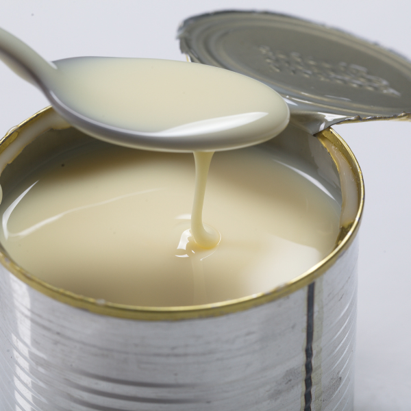 Open can of sweetened condensed milk with a spoon lifting the thick milk from the surface