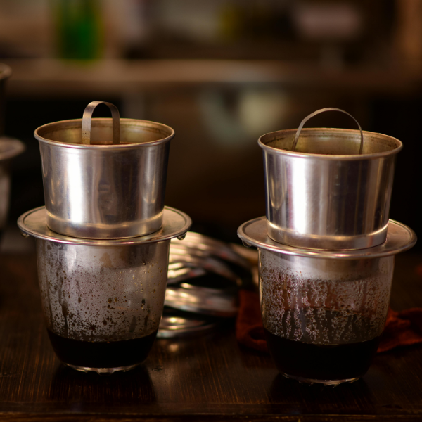 Two stainless steel Vietnamese phin filters dripping coffee into glass cups on a wooden table