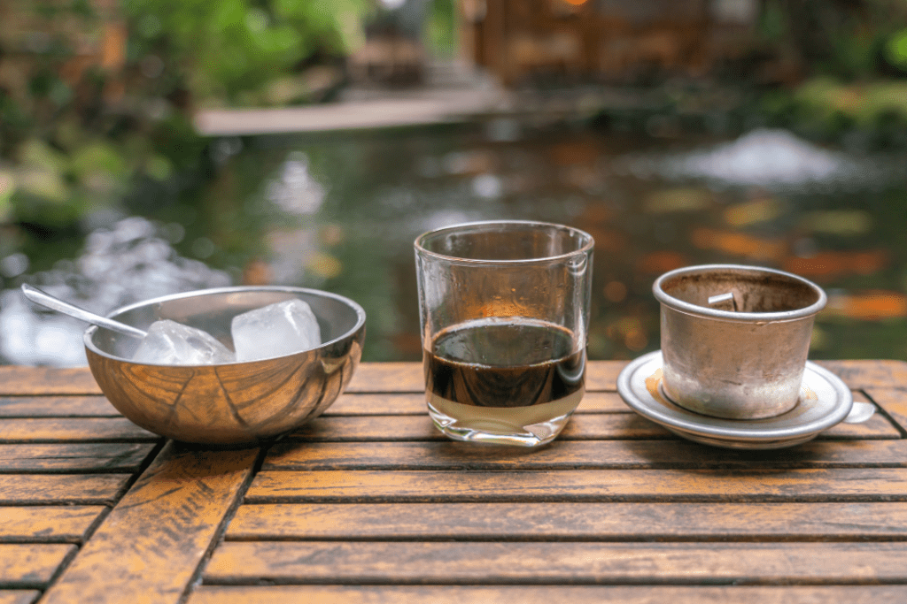 Glass of strong brewed coffee with condensed milk, phin filter, and bowl of ice on a wooden table outdoors
